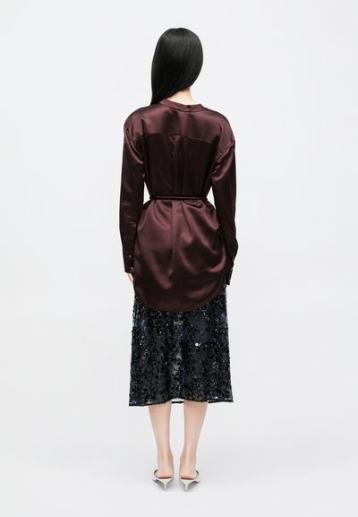 Γυναίκα με μακριά μαύρα μαλλιά, φοράει πουκάμισο μπορντό σατέν με ζώνη και μαύρη midi φούστα με παγιέτες, στέκεται με την πλάτη προς το μέρος μας, σε λευκό φόντο.