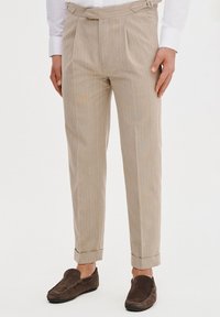 Beżowe spodnie w paski z zakładkami, z paskiem w talii i mankietami. Połączone z brązowymi butami typu slip-on. Gładka tekstura materiału.