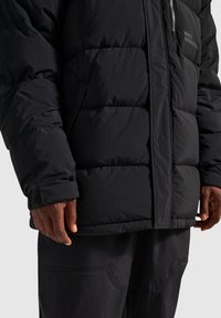 Sort isoleret nedjakke med kviltet design, lynlås foran og en syet lomme, båret over matchende mørke bukser.