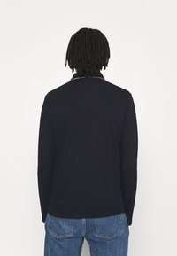 Person med axellång dreadlocks som bär en svart långärmad skjorta och blå jeans, stående vänd bort från kameran mot en vit bakgrund.