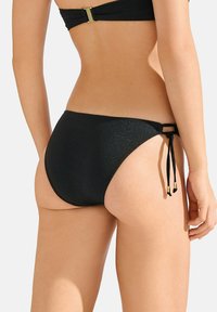 Zwarte bikini broek met een glanzende afwerking, zijdelings te strikken met gouden accenten, en een low-rise ontwerp voor minimale bedekking. Gladde textuur.
