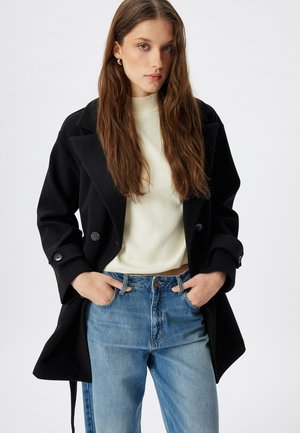 Cappotto di lana nero con due bottoni, ampi revers e tasche laterali, indossato sopra un maglione crema e jeans blu chiaro. Capelli lunghi e lisci.