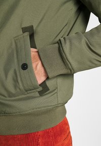 Main d’une personne dans la poche d’une veste vert olive avec poignets côtelés et bouton-pression noir, associée à un pantalon en velours côtelé rouge.