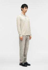 Μπεζ πόλο μπλούζα με μακριά μανίκια και γιακά, συνδυασμένη με ανοιχτό γκρι τζιν και μαύρα παπούτσια, στέκεται σε απλό φόντο.