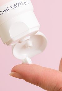 White cream is dispensed from a cream tube, resting on a fingertip. Soft pink background. Tube label shows 50 ml, 1.69 fl. oz.
