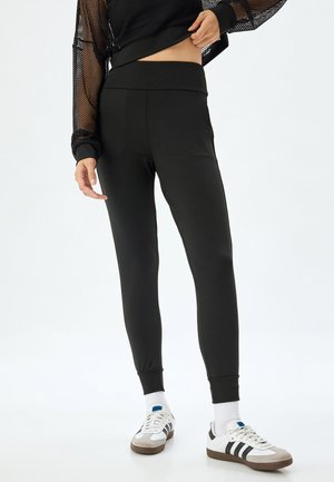 Black leggings with a wide waistband, smooth texture, and tapered legs. Worn with a black mesh top and white sneakers with gray accents.