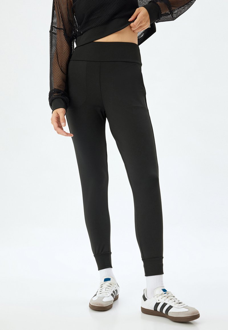 Sorte leggings med bred talje, glat tekstur og tapered ben. Båret med en sort nettop og hvide sneakers med grå detaljer.