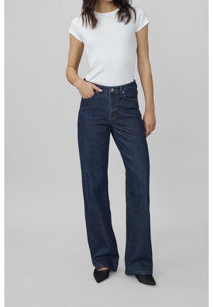 Femme portant un haut blanc ajusté à manches courtes et un jean large taille haute bleu foncé avec des chaussures noires à bout pointu, se tenant avec une main dans la poche.