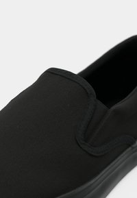 Gros plan d'une chaussure en toile noire à enfiler avec des panneaux latéraux élastiques et une semelle en caoutchouc sur un fond clair.