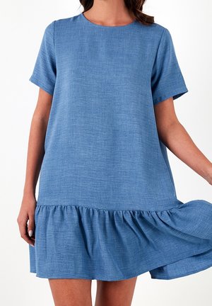 Mujer con vestido azul de manga corta y corte holgado con dobladillo con volantes, sosteniendo el vestido ligeramente hacia afuera sobre un fondo blanco.