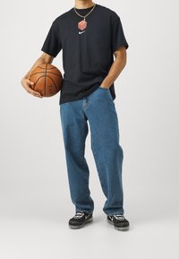Camiseta de algodón negra con el logotipo de Nike, jeans azules anchos y zapatillas negras con un diseño blanco. Sosteniendo un balón de baloncesto. Collar de cadena dorada.