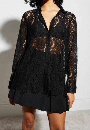Femme portant un chemisier en dentelle noire à manches longues avec des motifs floraux sur une jupe noire plissée, debout contre un mur blanc.