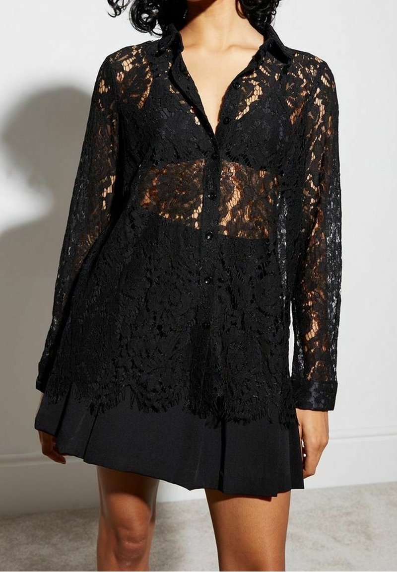 Femme portant un chemisier en dentelle noire à manches longues avec des motifs floraux sur une jupe noire plissée, debout contre un mur blanc.
