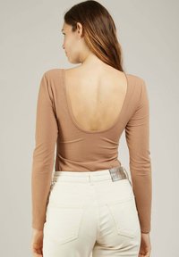 Body à manches longues, en maille côtelée, de couleur marron clair avec un décolleté plongeant au dos, associé à un jean blanc avec poches et une étiquette métallique brillante.