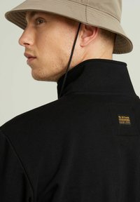 Chaqueta negra con cuello, de textura acanalada, con una etiqueta color caqui de G-Star en la parte posterior. El modelo lleva un sombrero de cubo beige.