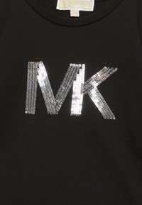 Černý textilní top se stříbrnými flitrovými písmeny "MK" umístěnými uprostřed pod límečkem, na kterém je bílým písmem vytištěno "Michael Kors".