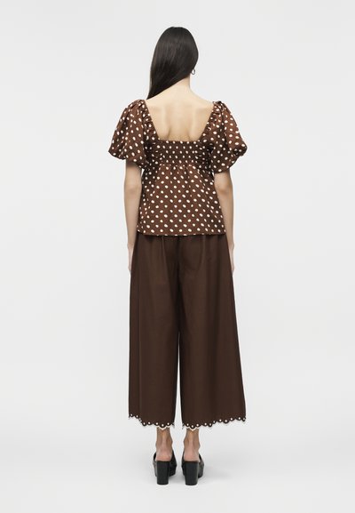 Γυναίκα με μακριά σκούρα μαλλιά που φοράει καφέ μπλούζα με λευκές πουά και φουσκωτά μανίκια, καθώς και καφέ παντελόνι φαρδύ με κυματιστά τελειώματα, στέκεται γυρισμένη προς τα πίσω.