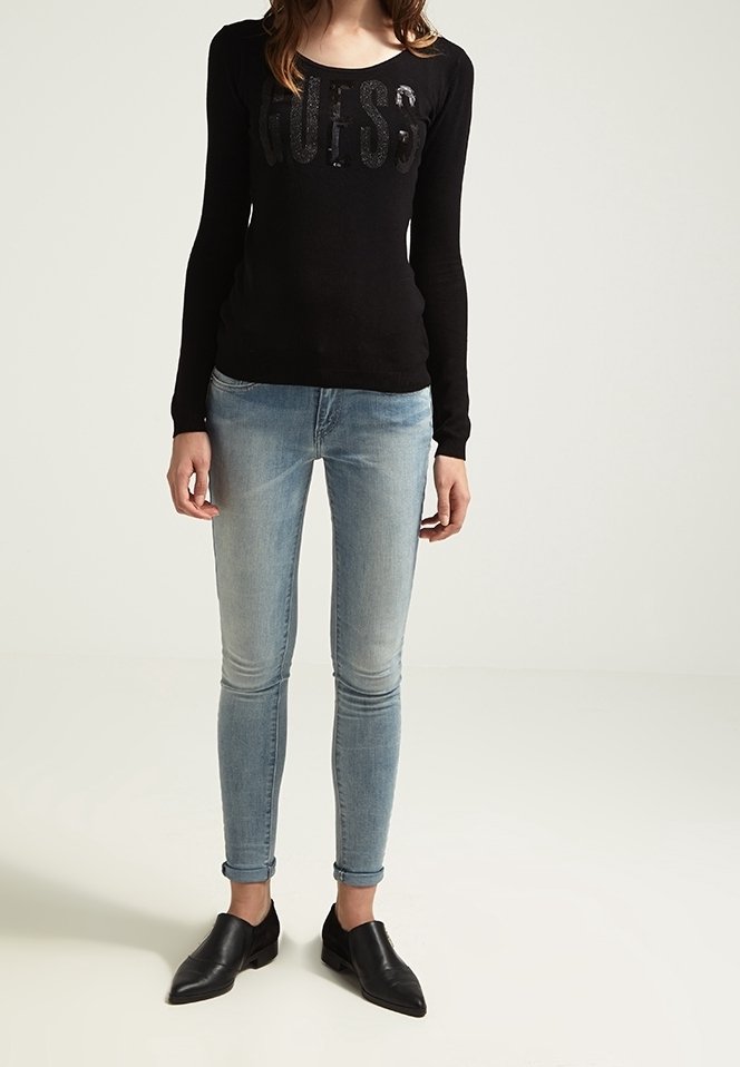 Femme portant une chemise noire à manches longues avec le logo "GUESS", un jean skinny bleu clair retroussé aux chevilles, et des chaussures noires à enfiler à bout pointu.