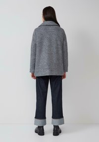 Femme portant une veste grise texturée, un jean large retroussé foncé et des bottines noires, se tenant debout contre un fond clair.