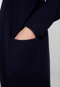 Mano parcialmente dentro del bolsillo delantero de un cárdigan de punto azul oscuro con mangas largas sobre un fondo claro.