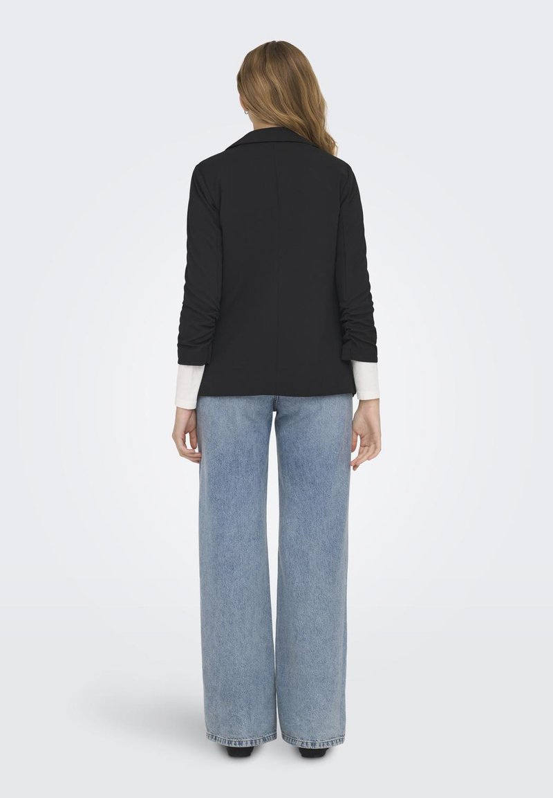 Zwarte blazer met verzamelde mouwen, gecombineerd met lichtblauwe wijde jeans. De blazer heeft een gladde textuur en een klassiek kraagontwerp.