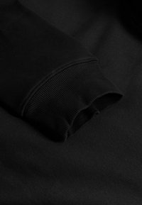 Juodas džemperis su raukšlėta rankogaliu. Medžiaga atrodo minkšta, lygaus paviršiaus, be matomų raštų ar metalinių detalių.