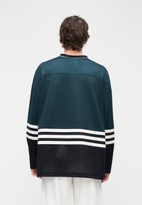 Hosszú ujjú mez sötét tengerzöld légáteresztő hálós anyagból, fehér vízszintes csíkokkal az alsó részen, fekete részletekkel és bordázott gallérral.