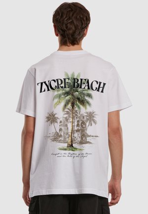Osoba nosząca białą koszulkę z nadrukiem palmy, głośników i napisu "Zycre Beach" na plecach.