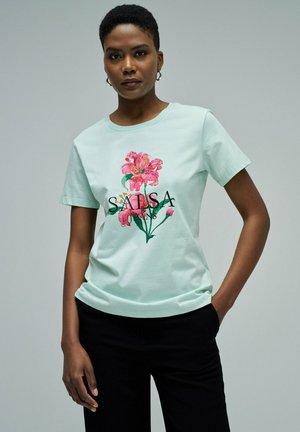 Mujer con el cabello corto, vestida con una camiseta azul claro con estampado floral rosa y pantalones negros, de pie con una mano en el bolsillo.