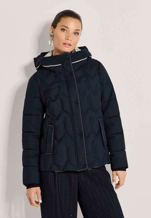 Frau, die eine schwarze gesteppte Kapuzenjacke mit Reißverschluss und Knopfverschluss sowie eine dunkle Nadelstreifenhose trägt, steht vor einer einfarbigen Wand.