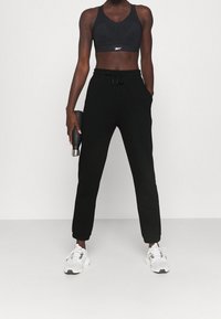 Svart sportbh och svarta joggerbyxor med fickor, avsmalnande ben och manschetter vid anklarna. Vita sneakers, håller en svart vattenflaska.