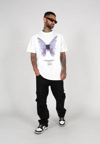Hvid t-shirt med et lila sommerfugl grafik og sort tekst. Kombineret med sorte cargo-bukser og sort-hvide sneakers.