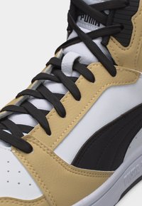 Κοντινό πλάνο σε αθλητικό παπούτσι Puma σε μπεζ, λευκό και μαύρο, που δείχνει μαύρα κορδόνια, ραμμένα δερμάτινα πάνελ και μέρος του λογοτύπου στη γλώσσα και στην σόλα.