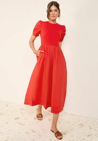 Robe midi rouge avec manches bouffantes, taille froncée et poches latérales. Texture lisse et tissu léger. Sandales à glisser brunes, fond en terrazzo.