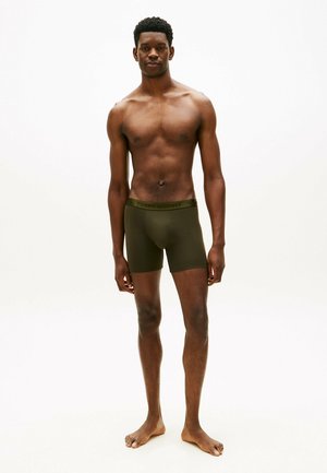 Jonge man staat op blote voeten en draagt olijfgroene boxershorts, kijkt naar voren tegen een effen witte achtergrond.