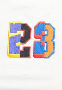 Λευκό ύφασμα με πολυχρωματισμένους κεντητούς αριθμούς "23" σε κίτρινο, μωβ, μαύρο, μπλε και πορτοκαλί με περιγραμμένα άκρα.