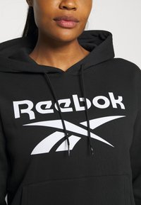 Czarna bluza z kapturem z dużym białym logo "Reebok" i graficznymi akcentami, z regulowanym sznurkiem i kieszenią z przodu.