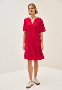 Kurzes rotes Kleid mit kurzen Ärmeln, V-Ausschnitt, gestuftem Design und strukturiertem Stoff. Kombiniert mit weißen Sneakers mit beigen Akzenten.