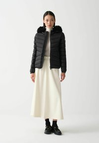 Schwarze Steppjacke mit einer Kapuze aus Kunstpelz, Reißverschluss und Rippbündchen, kombiniert mit einem langen crèmefarbenen Rock und schwarzen Loafers.