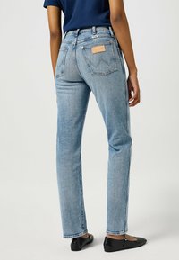 Jeans de mezclilla azul claro con corte recto, con bolsillos traseros y una etiqueta de cuero marrón. Combinados con zapatos planos negros, vistos desde atrás.