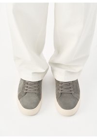 Baskets en daim gris avec semelles blanches portées avec un pantalon blanc ample sur fond blanc.