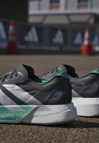 Coppia di scarpe da corsa grigie e verdi posizionate sull'asfalto con coni stradali arancioni e barriere sfocate sullo sfondo.