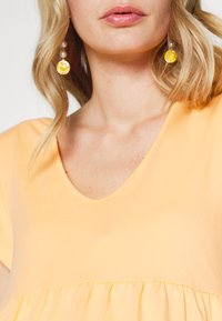 Top de algodão amarelo com um decote em V profundo e mangas curtas, apresentando uma parte inferior franzida. Os acessórios incluem brincos amarelos com o rosto sorridente.