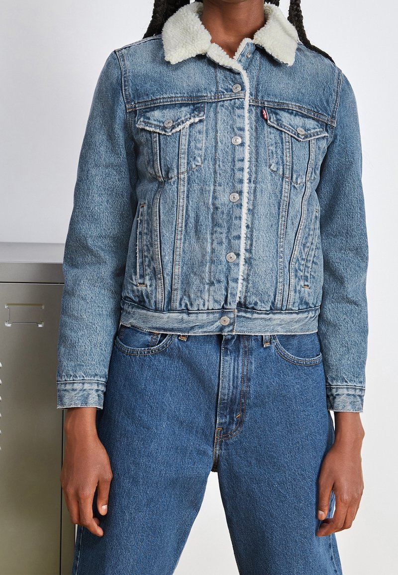 Veste en denim avec un délavage bleu clair, col en fausse fourrure, fermeture à boutons et poches poitrine, associée à un jean bleu taille haute.