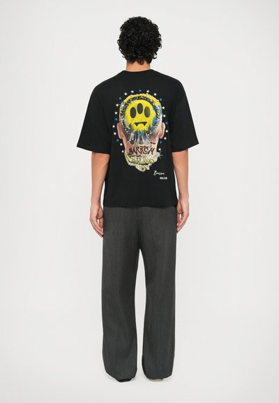 T-shirt nera oversize con una grafica colorata che presenta una faccina sorridente, stelle e testo sul retro. Pantaloni grigi a gamba larga.