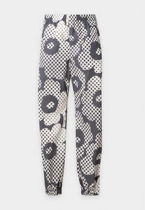 Homme debout les mains dans les poches, portant une chemise à manches courtes imprimée florale grise et blanche assortie avec un pantalon et des baskets marron.