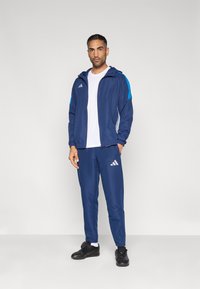 Σκούρο μπλε φόρμα με ανοιχτό μπλε λεπτομέρειες, που περιλαμβάνει ζακέτα με φερμουάρ και παντελόνι με ελαστική μέση, κατασκευασμένη από λείο, ελαφρύ ύφασμα.