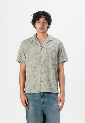 Chemise à manches courtes verte avec un motif de broderie florale, fermeture à boutons à l'avant et col classique, fabriquée à partir d'un tissu léger.