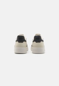 Baskets blanches avec un accent de talon noir, tige en cuir lisse, langue texturée et semelle en caoutchouc présentant des lignes horizontales sur le talon.