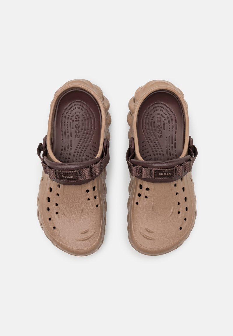 Zuecos ligeros, de color marrón, con correa ajustable y orificios de ventilación, que cuentan con plantillas texturizadas y bordes ondulados ligeramente elevados.